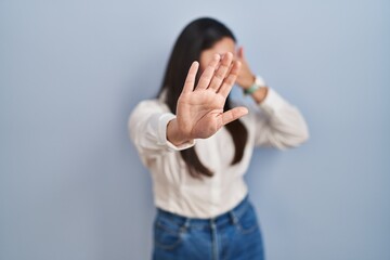 Young latin woman standing over blue background covering eyes with hands and doing stop gesture with sad and fear expression. embarrassed and negative concept.