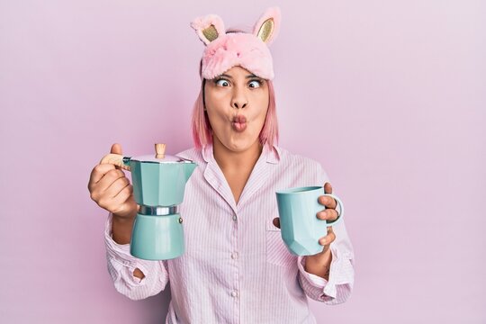 Hispanic woman with pink hair wearing sleep mask and pajama drinking a coffee cup making fish face with mouth and squinting eyes, crazy and comical.