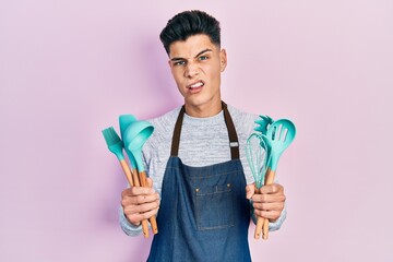 Young hispanic man wearing professional baker apron holding cooking tools clueless and confused...