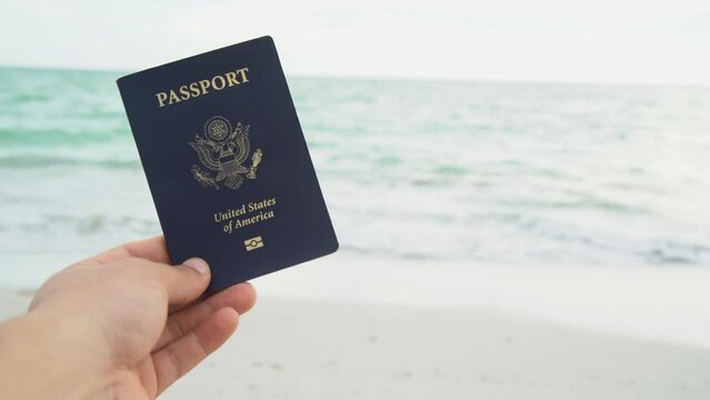 a hand holding two passport a red and a blue one on a beach with the waves of the sea in the background