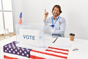 Handsome middle age man sitting at voting stand with a big smile on face, pointing with hand and finger to the side looking at the camera.