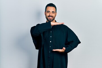 Handsome hispanic man with beard wearing catholic priest robe gesturing with hands showing big and...