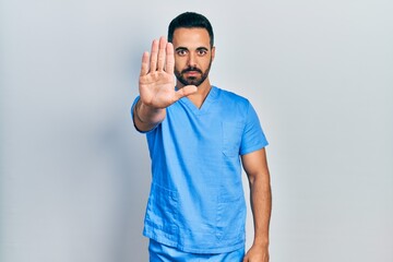 Handsome hispanic man with beard wearing blue male nurse uniform doing stop sing with palm of the...