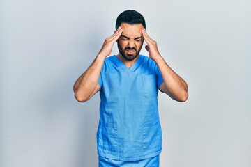 Handsome hispanic man with beard wearing blue male nurse uniform with hand on head for pain in head because stress. suffering migraine.