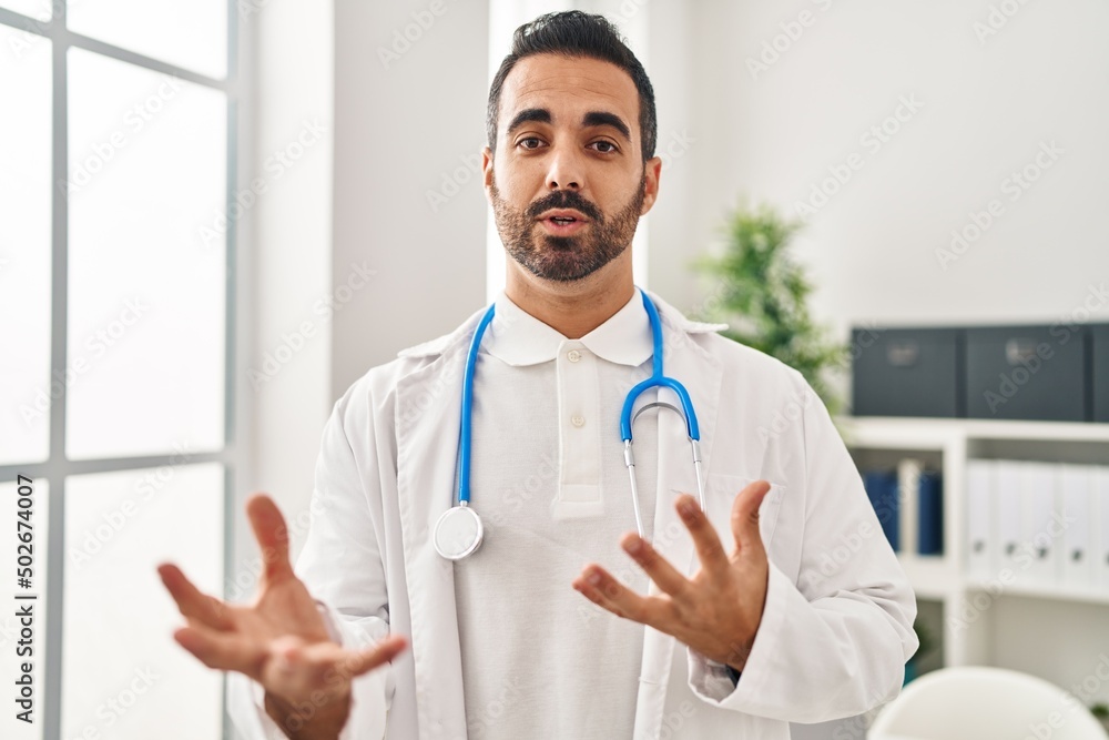 Sticker Young hispanic man wearing doctor uniform speaking at clinic