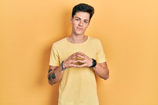 Young hispanic man wearing casual yellow t shirt hands together and fingers crossed smiling relaxed and cheerful. success and optimistic