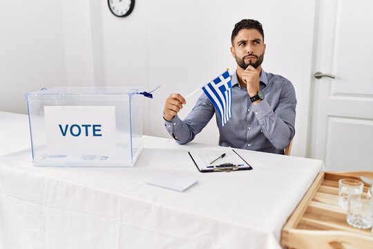 Young handsome man with beard at political campaign election holding greece flag serious face thinking about question with hand on chin, thoughtful about confusing idea