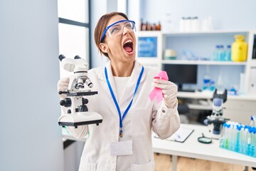 Young brunette woman working at scientist laboratory holding pink ribbon angry and mad screaming frustrated and furious, shouting with anger looking up.