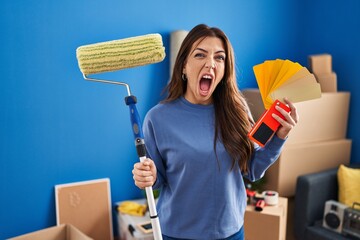 Young brunette woman holding roller painter painting new house angry and mad screaming frustrated...