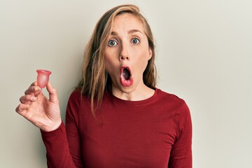 Young blonde woman holding menstrual cup scared and amazed with open mouth for surprise, disbelief...