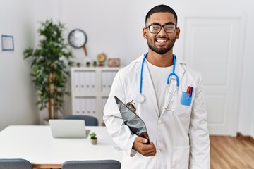 Young indian man wearing doctor uniform and stethoscope winking looking at the camera with sexy expression, cheerful and happy face.