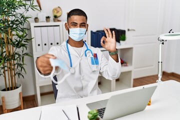 Young indian doctor offering safety mask doing ok sign with fingers, smiling friendly gesturing excellent symbol