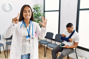 Young asian doctor woman at waiting room with a man with a broken arm showing and pointing up with fingers number six while smiling confident and happy.
