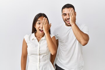 Young interracial couple standing together in love over isolated background covering one eye with hand, confident smile on face and surprise emotion.