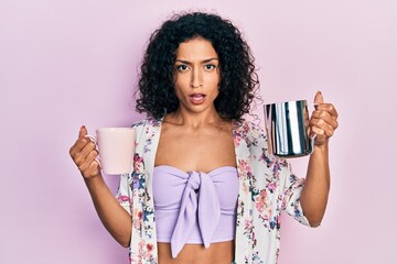 Young latin girl holding cup of coffee and jar of milk in shock face, looking skeptical and...