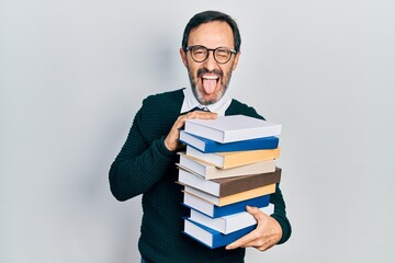 Middle age hispanic man holding a pile of books sticking tongue out happy with funny expression.