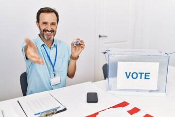 Middle age man with beard sitting by ballot holding i vote badge smiling friendly offering...