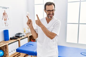 Obraz premium Middle age man with beard working at pain recovery clinic smiling and looking at the camera pointing with two hands and fingers to the side.