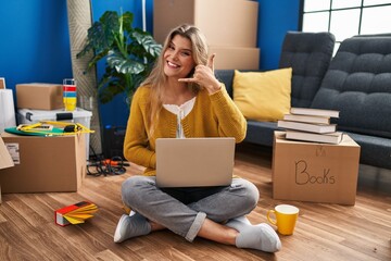 Young woman sitting on the floor at new home using laptop smiling doing phone gesture with hand and fingers like talking on the telephone. communicating concepts.