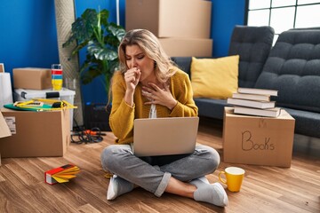 Young woman sitting on the floor at new home using laptop feeling unwell and coughing as symptom...