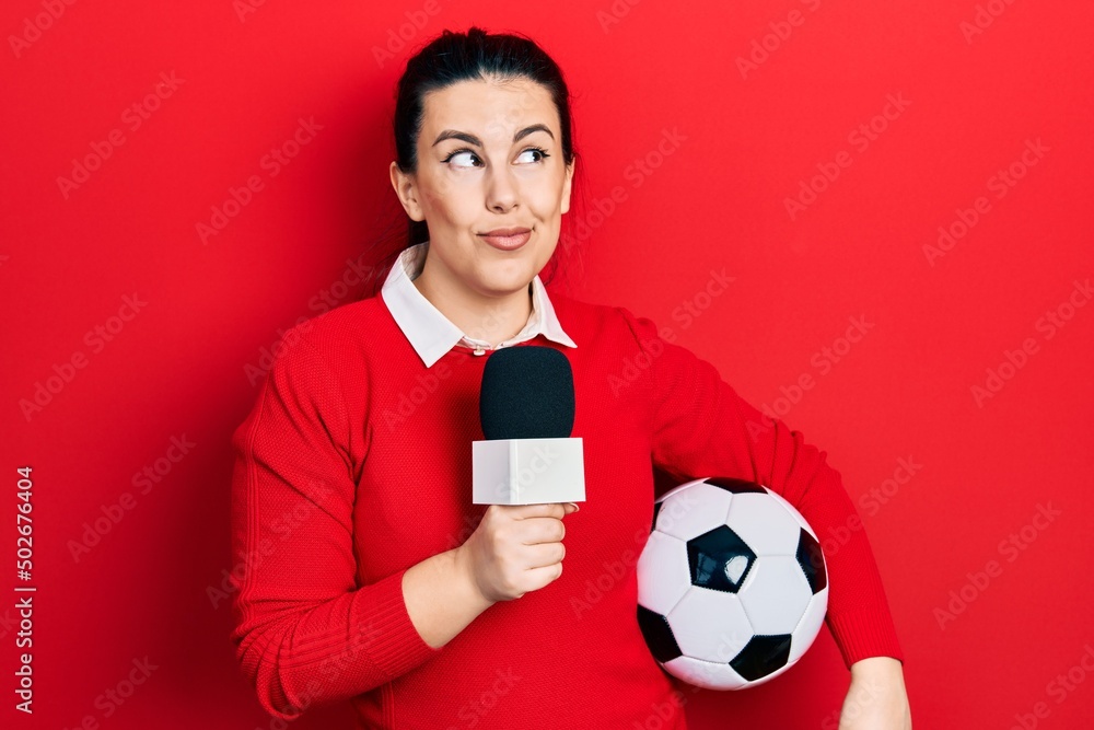 Sticker Young hispanic woman holding reporter microphone and soccer ball smiling looking to the side and staring away thinking.