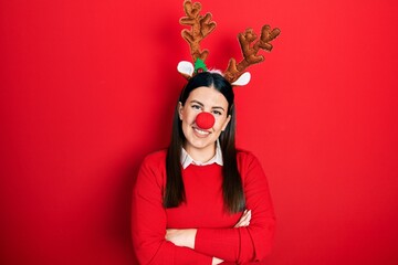 Young hispanic woman wearing deer christmas hat and red nose happy face smiling with crossed arms...