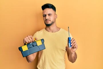 Young arab man holding toolbox and screwdriver clueless and confused expression. doubt concept.