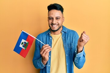 Young arab man holding haiti flag screaming proud, celebrating victory and success very excited with raised arm - obrazy, fototapety, plakaty