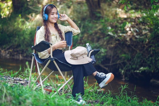 A young woman sits relaxing by the forest river and wears headphones to listen to music happily.