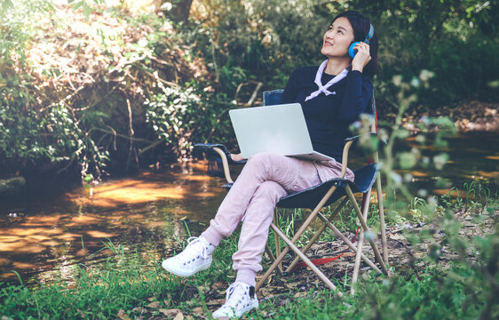 A young woman sits and relaxes by the forest river and wears headphones to listen to music happily.