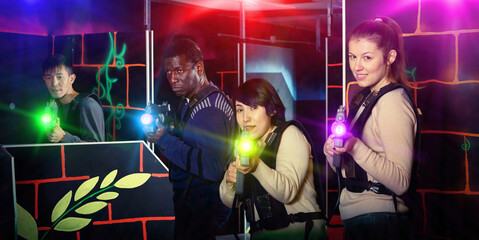 Modern cheerful smiling people of different nationalities with laser pistols playing laser tag on dark labyrinth
