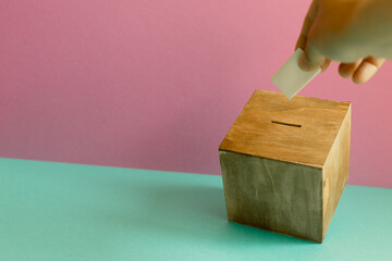 Hand putting voting paper in Ballot box on mint green table. pink wall background