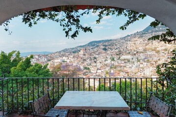 Behind the balcony of the hotel is a city on the slopes of the mountains. The rays of the morning sun and the roofs of houses. Colonial style chairs and table for relaxing in Taxco, Mexico