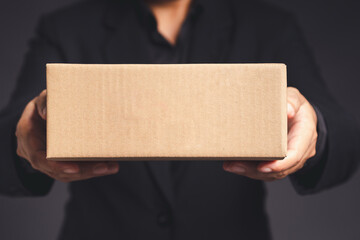 Businessman in a black suit holds and points to a cardboard box while standing on a gray background