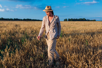 Naklejka na ściany i meble Tall handsome man dressed in a coarse linen suit and hat standing at golden oat field