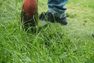Close-up shot of male worker in jeans maintenance garden using an electric lawnmower to trim overgrown green grass in the lawn yard, scattering the grass in summer, outdoor landscaping work service.