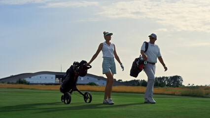 Two golfers walk fairway at sunset. Rich couple carry sport equipment clubs.