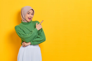 Shocked beautiful Asian woman in green sweater pointing fingers aside at copy space isolated over yellow background