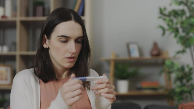Caucasian scared woman checking positive result on predictor test while sitting on sofa in living room. Unwanted pregnancy and unplanned motherhood concept.