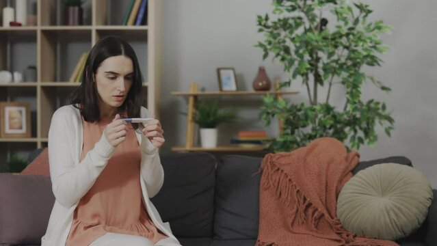 Caucasian young woman sitting alone on couch and feeling shocked with positive unplanned pregnancy test result. Hard decision and unwilling pregnancy concept.