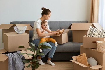 Portrait of smiling dark haired woman wearing white t shirt sitting on sofa surrounded with carton...