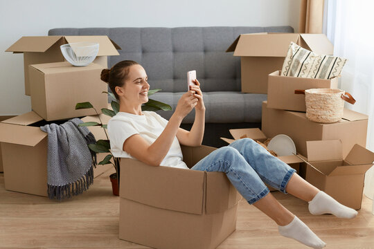 Side view of positive optimistic woman wearing white t shirt and jeans sitting in a cardboard box and using smart phone, having rest while unpacking her belongings, typing messages.