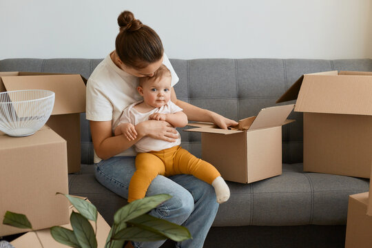 Portrait of woman with bun hairstyle wearing white t shirt sitting on sofa and holding her infant baby, unpacking boxes together with her daughter, young family moving into a new apartment.