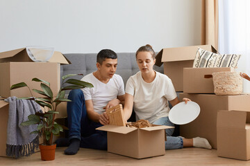 Indoor shot of shocked couple relocated in a new apartment and unpacking cardboard boxes with...