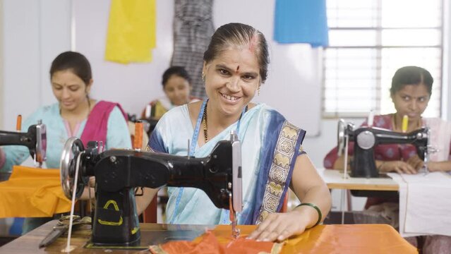Happy smiling woman showing thumbs up sign by looking camera while working at garments sewing maching - concept of employment, job and successful business