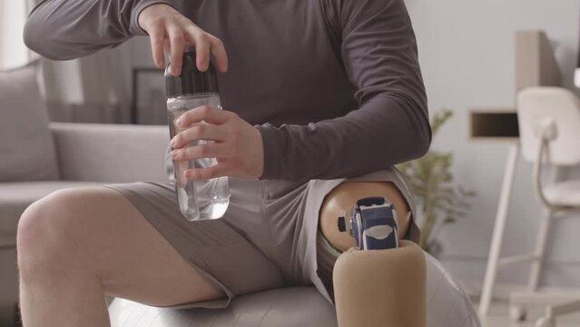 Tilt up slowmo of young Caucasian man with prosthetic leg drinking water while sitting on git ball having home workout
