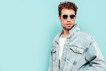 Portrait of handsome confident stylish hipster lambersexual model with curly hairstyle. Sexy man dressed in jeans jacket. Fashion male isolated on blue wall in studio. In sunglasses