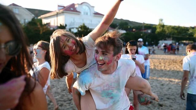 Smiling man carries pretty woman on back both stained in colored powder at Holi festival on beach. Couple has fun with friends at hindu holiday of colors, love, unity. End of covid pandemic isolation