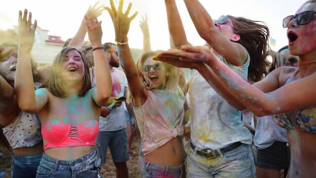 Shouting women smeared in colorful powder throw dry colors up in air to celebrate Holi festival on beach in slow motion. Outdoor hindu holiday party. End of lockdown, covid pandemic, restrictions.