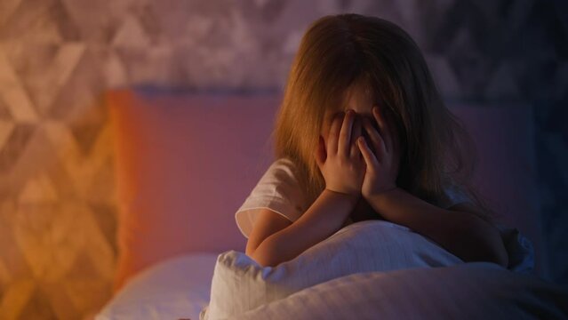 Unhappy child hides closing eyes with palms on bed in room